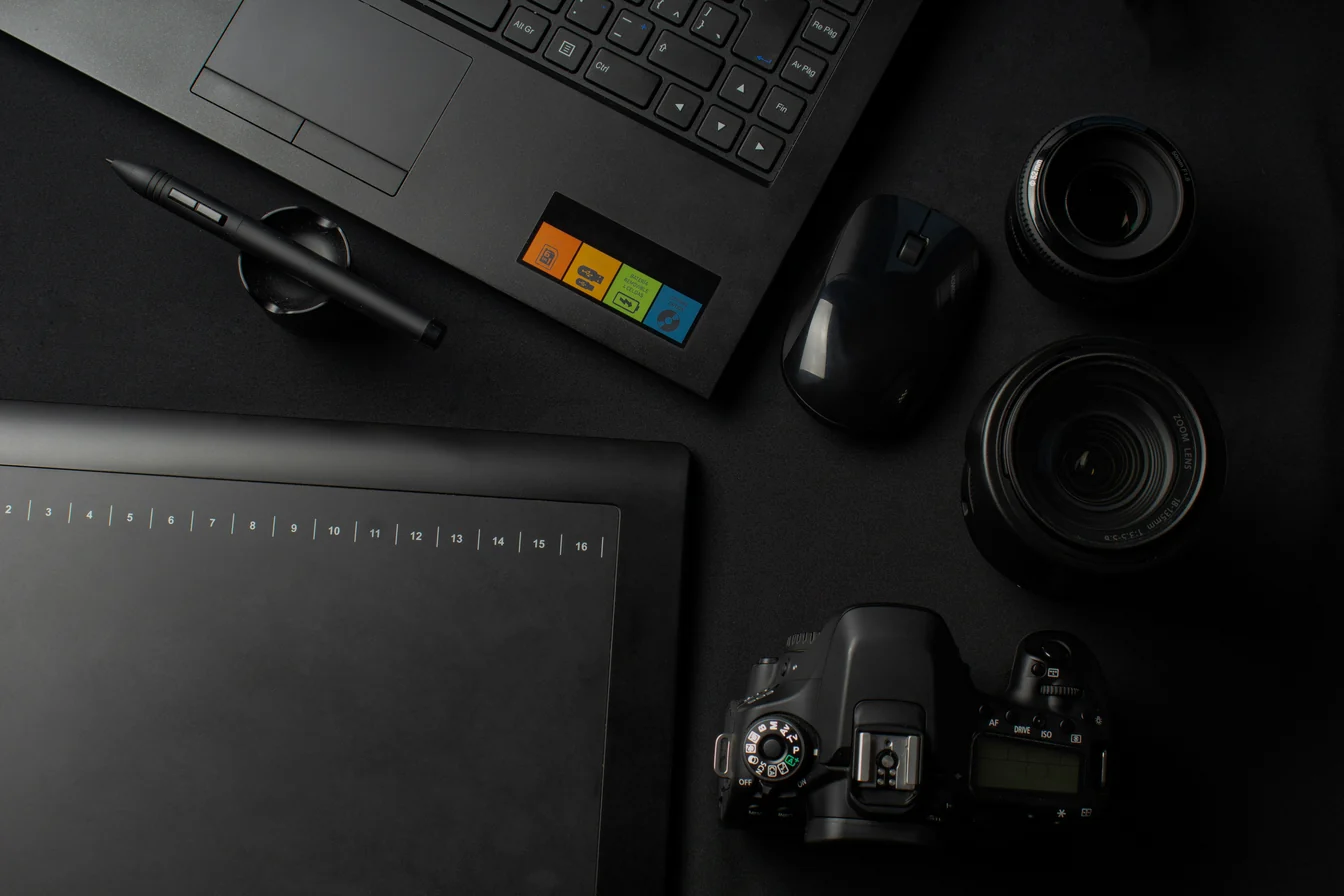 Overhead view of a dark desk with a laptop, graphics tablet and stylus, mouse, camera lenses and a DSLR camera.